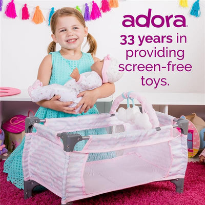 A young girl in a teal dress smiles while holding a doll, next to a pink and gray pack-n-play by Adora.