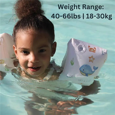 Child wearing Ali + Oli inflatable swim arm band wings in a pool, with visible sea creature designs and safety weight range.