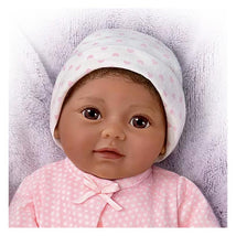 Lifelike doll named Kiara, wearing a pink polka-dot outfit and a white hat, resting on a soft blanket.