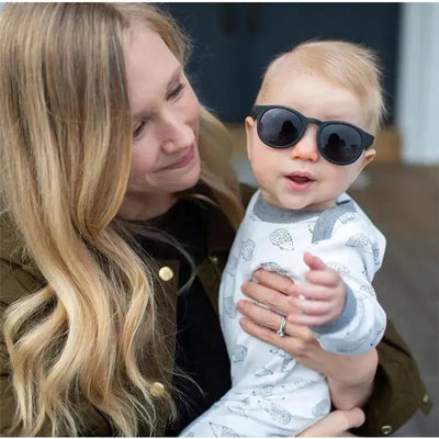 Baby wearing black Babiators sunglasses, being held by a smiling woman, both appearing happy in a warm outdoor setting.