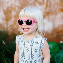 A smiling toddler with short blonde hair wears bright pink Babiators sunglasses, standing in front of greenery.