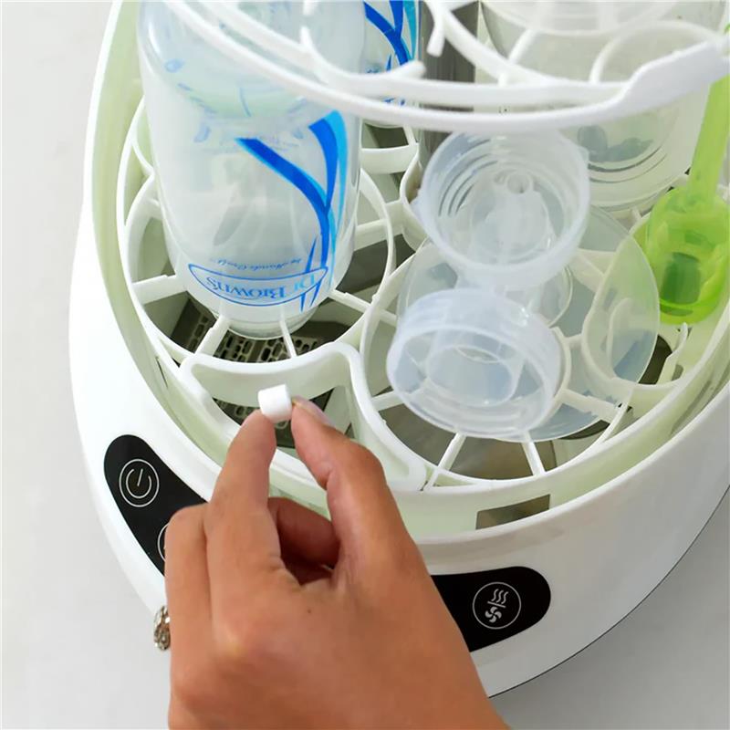 Hand holding a Baby Brezza soap tablet above a bottle washer filled with baby bottles and accessories.