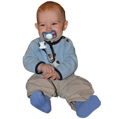 A smiling baby with a blue pacifier sits on the floor, wearing a light blue sweater and beige pants with blue socks.