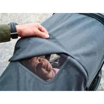 A hand lifts the canopy of the Baby Jogger City Prix™ Jogger X Bike Trailer, revealing a curious child inside.