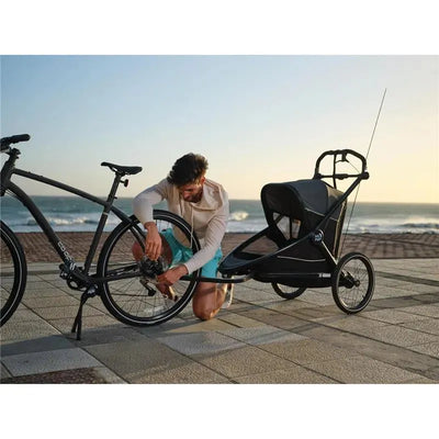 A person in a light hoodie is kneeling and attaching the Baby Jogger City Prix™ Trailer Conversion Kit to a black bicycle near the beach.