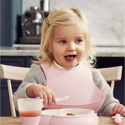 Toddler wearing a Babybjorn powder pink bib while eating at a table, smiling, with a cup and bowl in front.