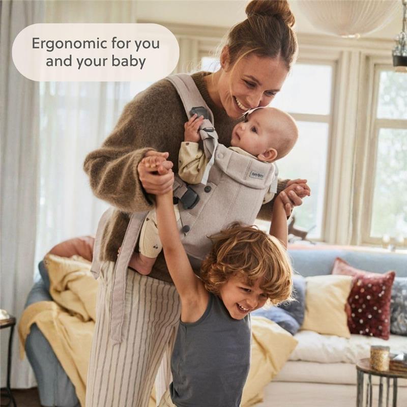 Mom holds her baby in a light beige BabyBjörn Baby Carrier while lifting her smiling child in a cozy living room.
