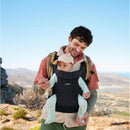 A man smiling holds a baby in a Babybjorn Baby Carrier in Sand Gray, outdoors with a scenic landscape in the background.