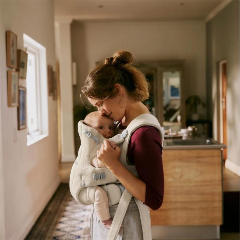 Mother using a Babybjorn Baby Carrier in Sand Gray, gently kissing her baby while standing in a cozy indoor space.
