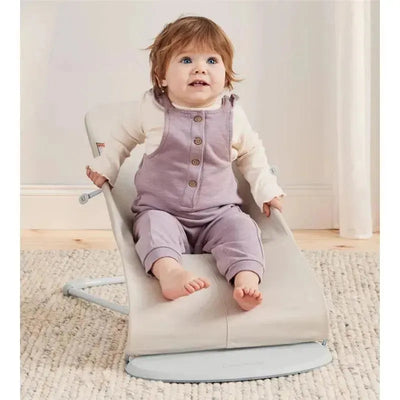 Light beige BabyBjorn Bouncer Bliss with a smiling baby in a purple outfit, sitting comfortably on the bouncer.
