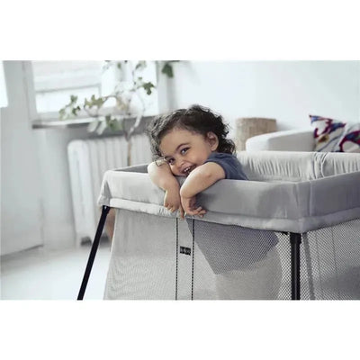 Toddler smiling and leaning on the side of a BABYBJORN Travel Crib Light in silver with a fitted sheet.