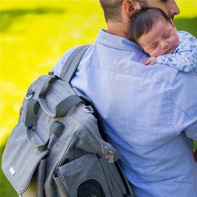 Charcoal Bbluv Metrö Complete Backpack Diaper Bag slung over the shoulder of a person holding a sleeping baby.
