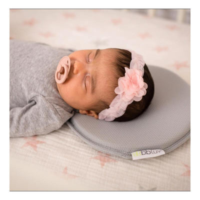 Baby resting on a gray Bblüv Pilo ergonomic headrest, wearing a pink flower headband and pacifier, on a patterned crib sheet.