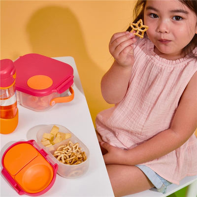 A young girl in a pink dress holds a star-shaped pretzel while seated at a table with colorful snack containers and a water bottle.