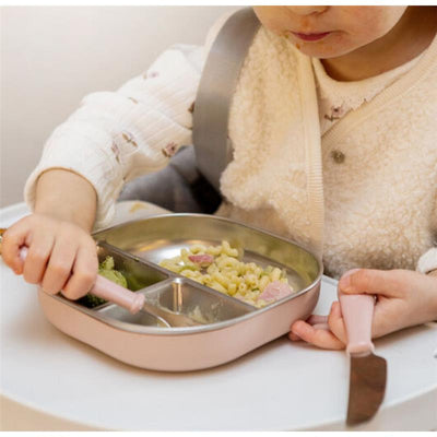 A toddler uses a pink Beaba 3Pk stainless steel cutlery set to eat pasta and veggies from a divided plate.