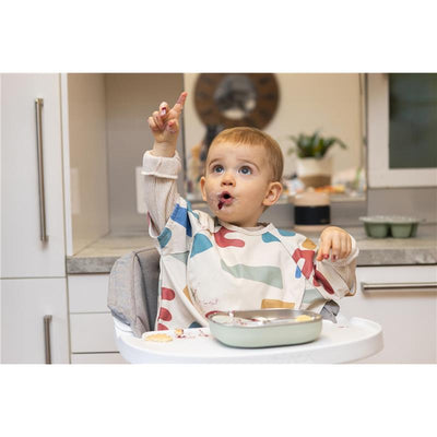 A toddler in a colorful bib sitting in a high chair, pointing excitedly, with a Beaba stainless steel divided plate in front.