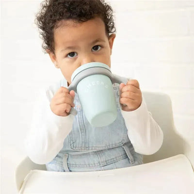 Light blue Bella Tunno Happy Sippy Cup held by a child, who is sipping from it while seated at a table.