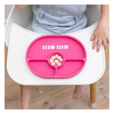 Bright pink Bella Tunno Miss Mess Wonder Plate on a white high chair table, holding small pieces of food, with a child’s hand nearby.