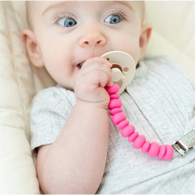 Baby holding a pink Bella Tunno Punch Pacifier Clip attached to a pacifier, sitting in a crib.