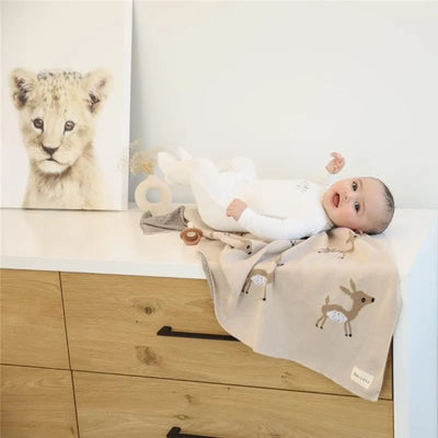 Luxury cotton baby swaddle blanket featuring deer design, draped beside a smiling baby on a dresser with a lion cub painting.