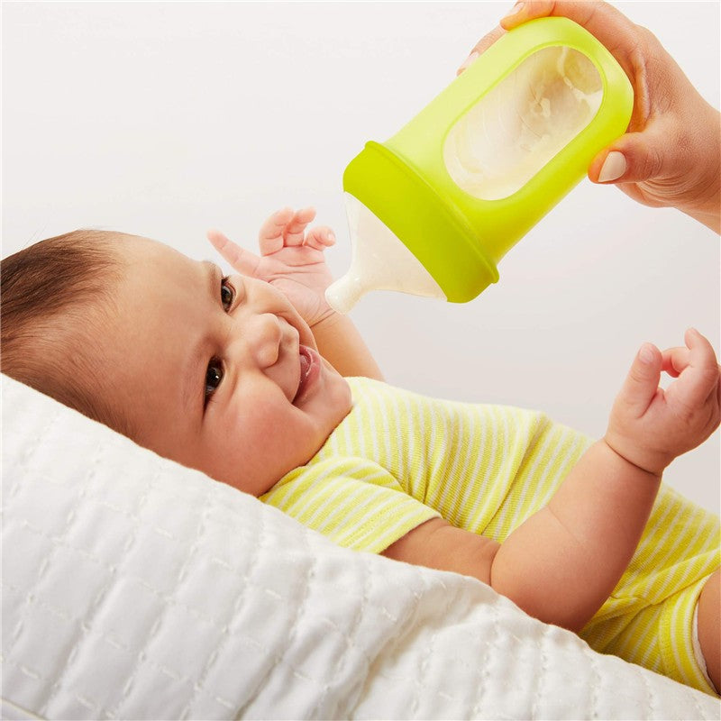 A smiling baby in a striped yellow onesie being fed from a Boon NURSH silicone bottle nipple in a green holder.