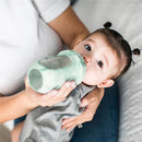 Baby with pigtails drinking from a mint tie-dye Boon Nursh 8 oz baby bottle, held by an adult.