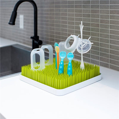 Boon TWIG drying rack accessories in white and gray, featuring colorful utensils and bottles on a green grass-like base.