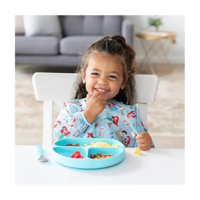 Little girl wearing a Disney-themed sleeved bib, smiling while eating from a divided blue plate with food.