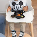 Child in a high chair wearing a Mickey Mouse bib, using a black silicone feeding set with mashed food in bowls.