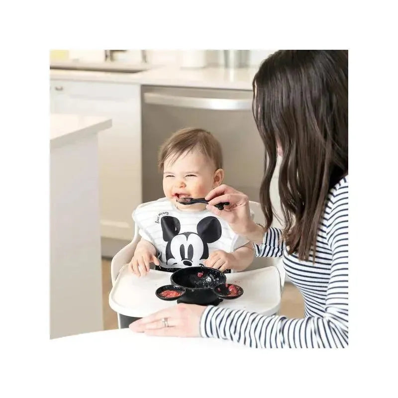 Baby wearing a Mickey Mouse shirt smiles as an adult feeds them with a spoon using a Bumkins Disney silicone feeding set.