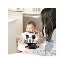 Baby wearing a Mickey Mouse shirt smiles as an adult feeds them with a spoon using a Bumkins Disney silicone feeding set.