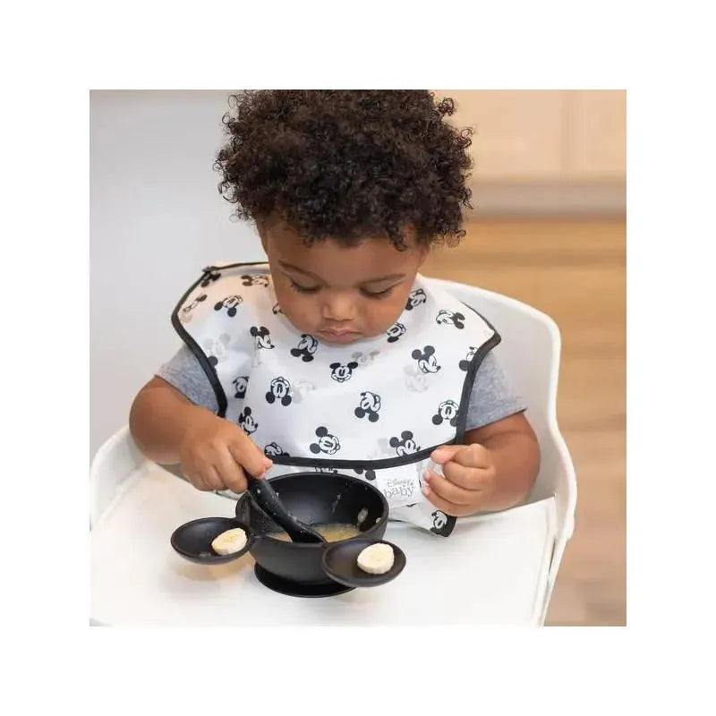 Child wearing a Mickey Mouse bib using a spoon to eat from a black silicone bowl with a small scoop attached.