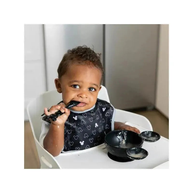 Toddler in a high chair wearing a black Mickey Mouse bib, holding a spoon and sitting next to a black feeding bowl.