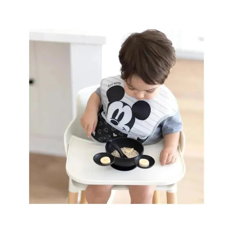 Toddler wearing a Mickey Mouse bib sits in a high chair, using a black silicone feeding set with small bowls and spoon.