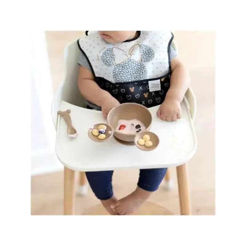 Baby wearing a Mickey Mouse bib sits in a high chair with a silicone feeding set, including bowls of food and a spoon.