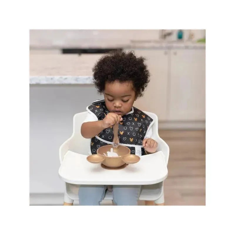 Toddler in high chair using a spoon to eat from a gold silicone feeding bowl set featuring Disney designs.