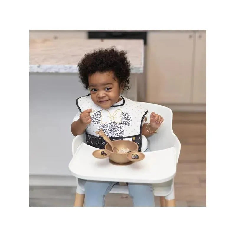 Toddler wearing a Disney-themed bib sits in a high chair with a bowl and spoon, smiling.