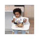 Toddler wearing a Disney-themed bib sits in a high chair with a bowl and spoon, smiling.