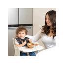 A toddler with curly hair in a high chair, wearing a Disney-themed bib, interacts with a caregiver over a feeding bowl.
