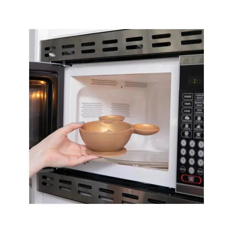 A hand holding a gold Bumkins Disney silicone feeding bowl set inside a microwave.