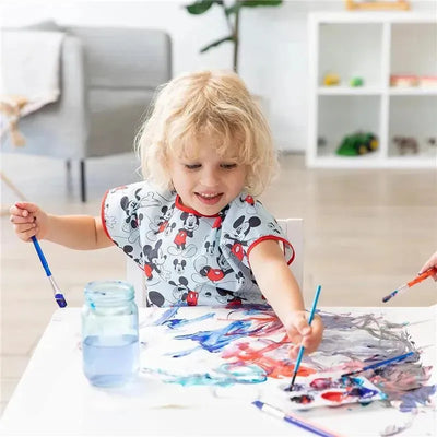 Child wearing a Mickey Mouse-themed smock, happily painting on paper with blue and red paint, surrounded by art supplies.