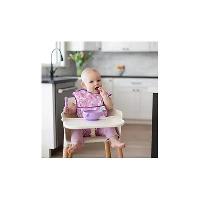 A baby in a high chair wearing a purple bib is enjoying food with a lavender silicone bowl and spoon.