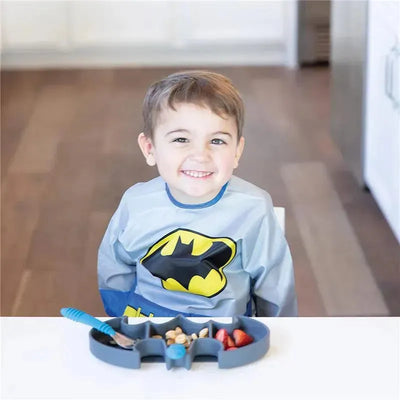 Child wearing a Batman-themed bib sits at a table with a silicone Batman-shaped dish filled with snacks.