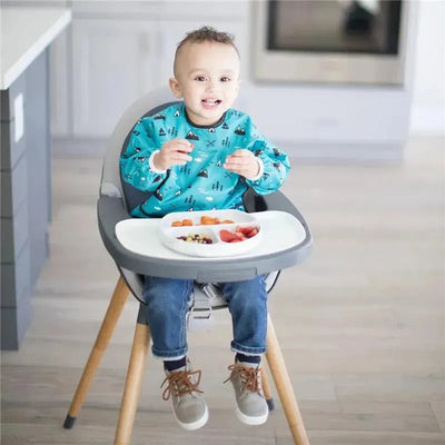 A smiling child in a high chair wearing a blue shirt with patterns is enjoying food from a divided silicone dish.