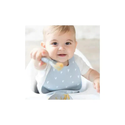Baby wearing a light blue silicone bib with white hearts, happily holding a spoon with food while seated in a high chair.