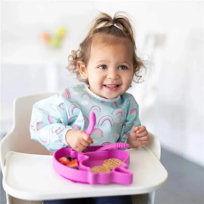 Toddler wearing a Bumkins sleeved bib with rainbow designs, happily eating from a pink plate containing fruit and snacks.