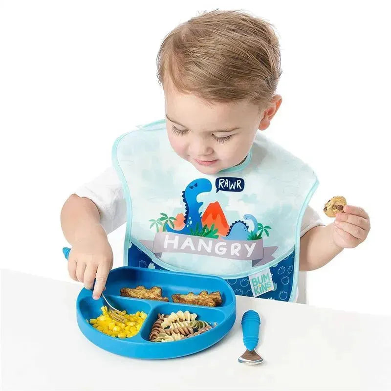A young child in a dinosaur-themed bib is enjoying a meal with a blue Bumkins spoon and fork, featuring pasta, corn, and snacks.