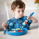 Child using a dark blue Bumkins spoon and fork set to eat from a divided blue plate with food.