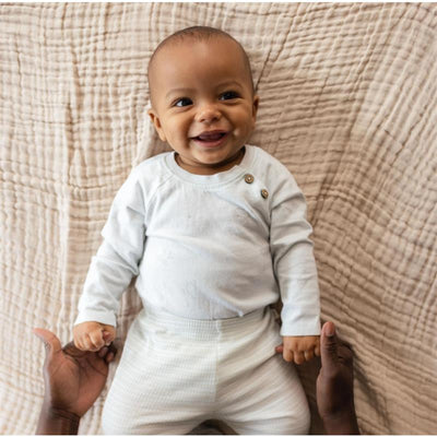 Smiling baby wearing a light blue long-sleeve bodysuit with button detail and light patterned pants, held by adult hands.