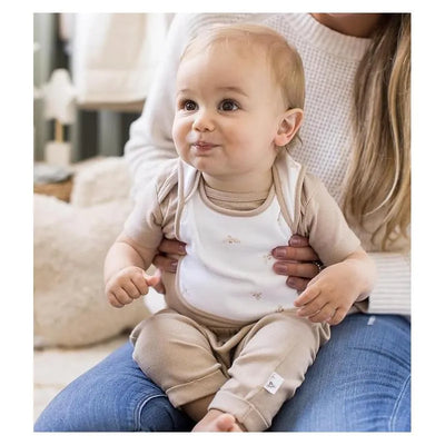 Baby girl in beige and white bodysuit with floral pattern, sitting on a lap, smiling, captured indoors.